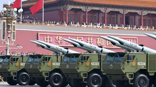 A series of green tanks holding large missiles roll on the streets below a large red building where people are standing next to China's national flags. 